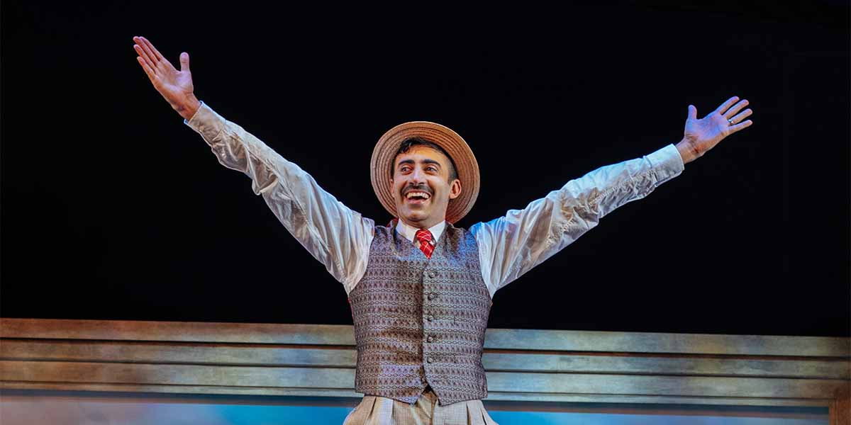 Richard David-Caine, in a straw hat and waistcoat, joyfully raises his arms on stage.