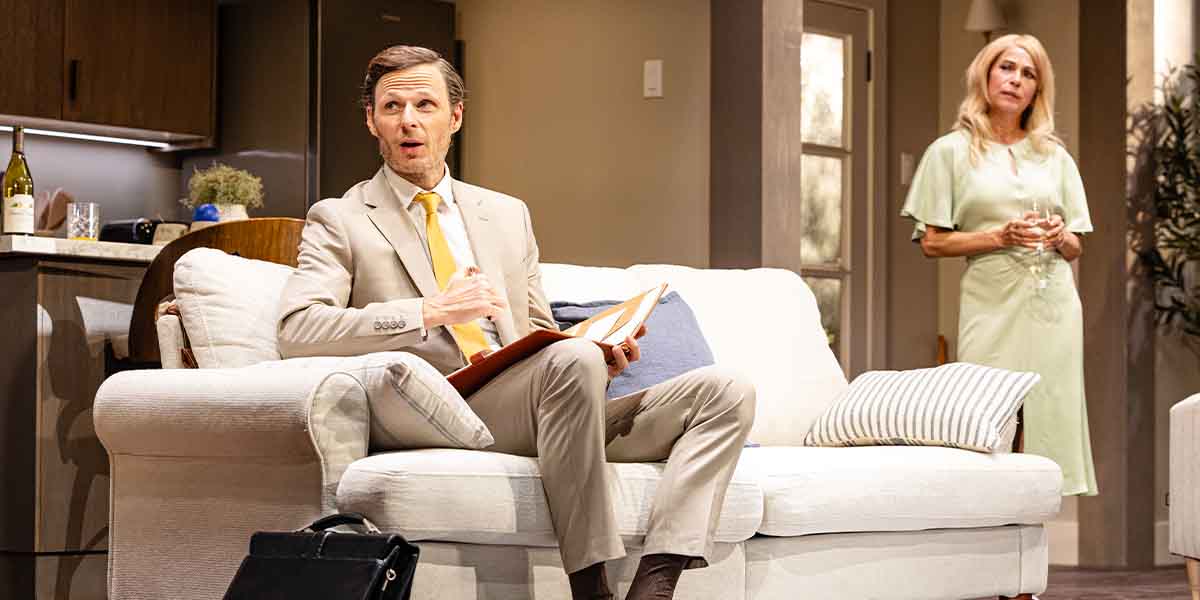 Rupert Young as Brad sits on a sofa, holding a notebook, while Juliet Cowan as Ellen stands nearby.