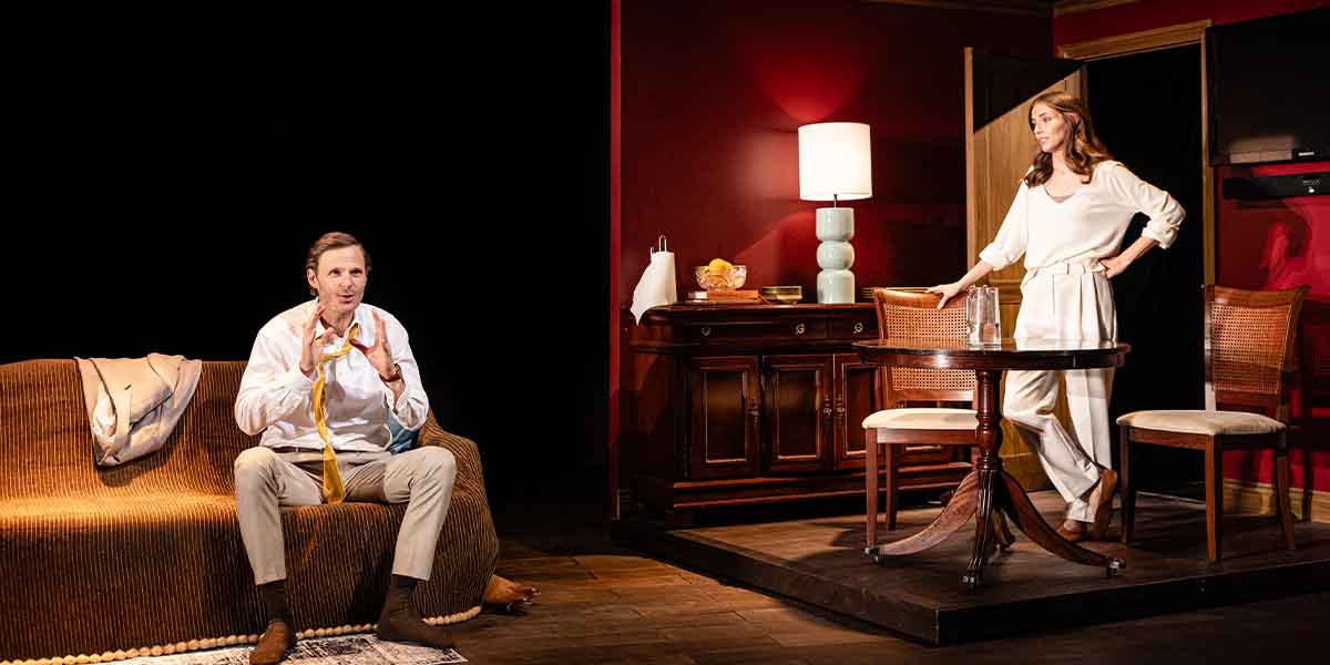 Rupert Young and Wallis Currie-Wood perform in 'Affluenza', seated in a stylish, dimly lit set.