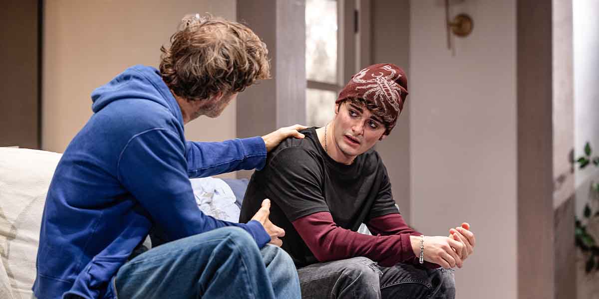 Rupert Young and Noah Schnapp sit on a sofa, engaged in a serious conversation, indoors.