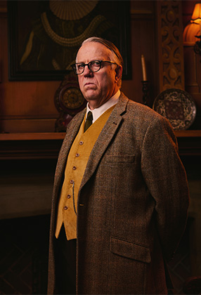 A man in glasses and a tweed coat stands confidently, with a yellow vest, against a wooden backdrop.