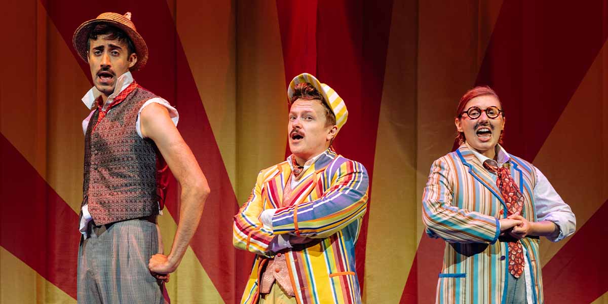 Richard David-Caine, Jonny Weldon, and Bronté Barbé perform in colourful costumes against a vibrant backdrop.
