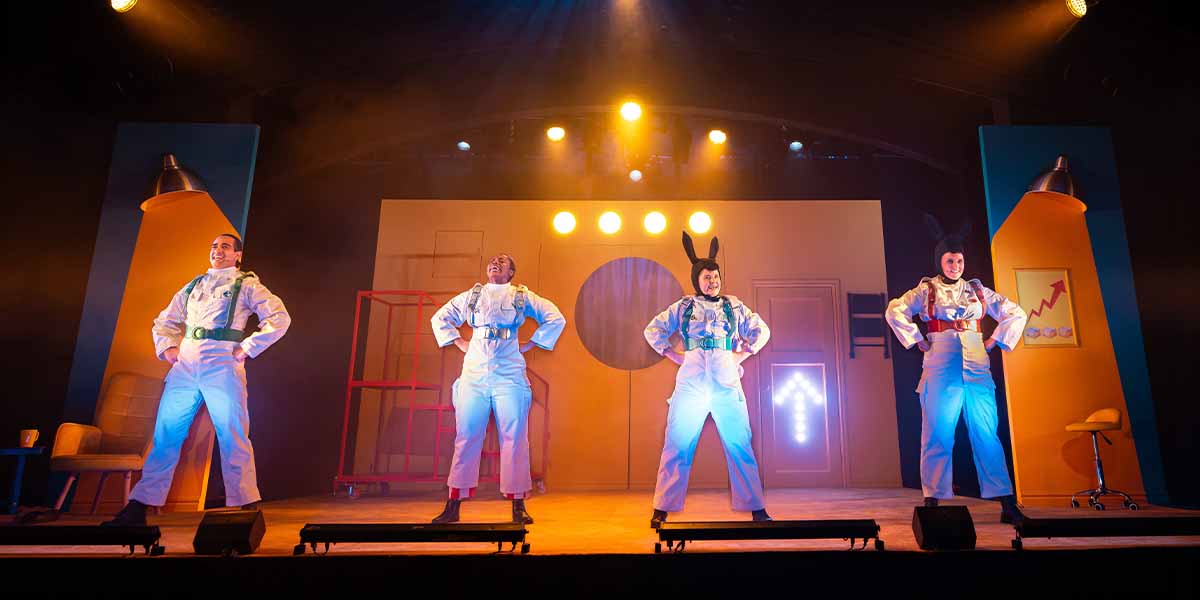 Four performers in white costumes pose confidently on stage, with colourful lighting and a simple backdrop.