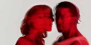 A woman and a man are closely positioned, their faces blurred with a red hue against a light background.