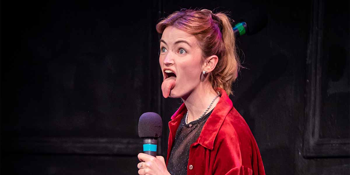 Jade Oswald sticks out her tongue while holding a microphone, wearing a red jacket against a dark background.