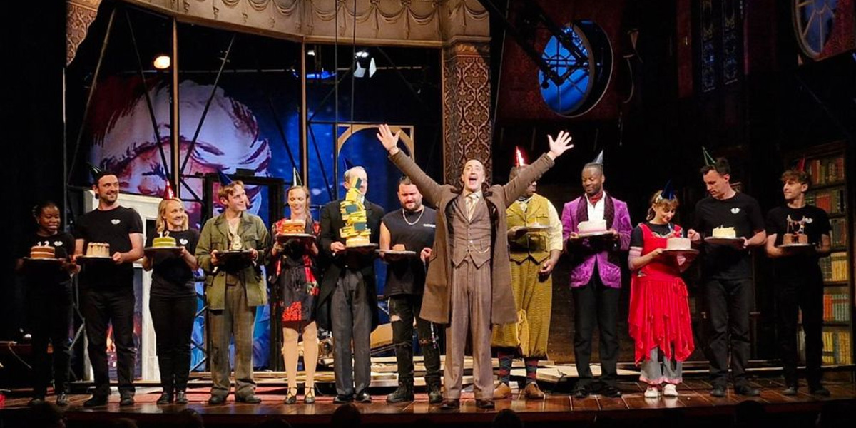 Cast members of The Play That Goes Wrong celebrate its 12th birthday with a cake and balloons.