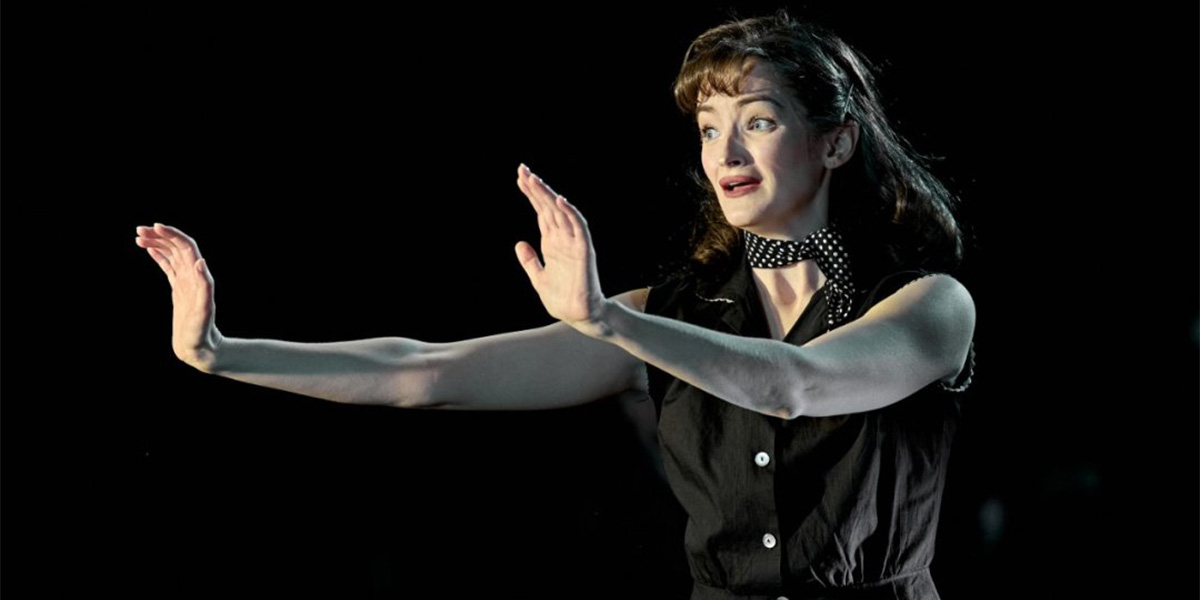 Jade Oswald performs expressively, wearing a black dress and polka dot scarf, against a dark background.