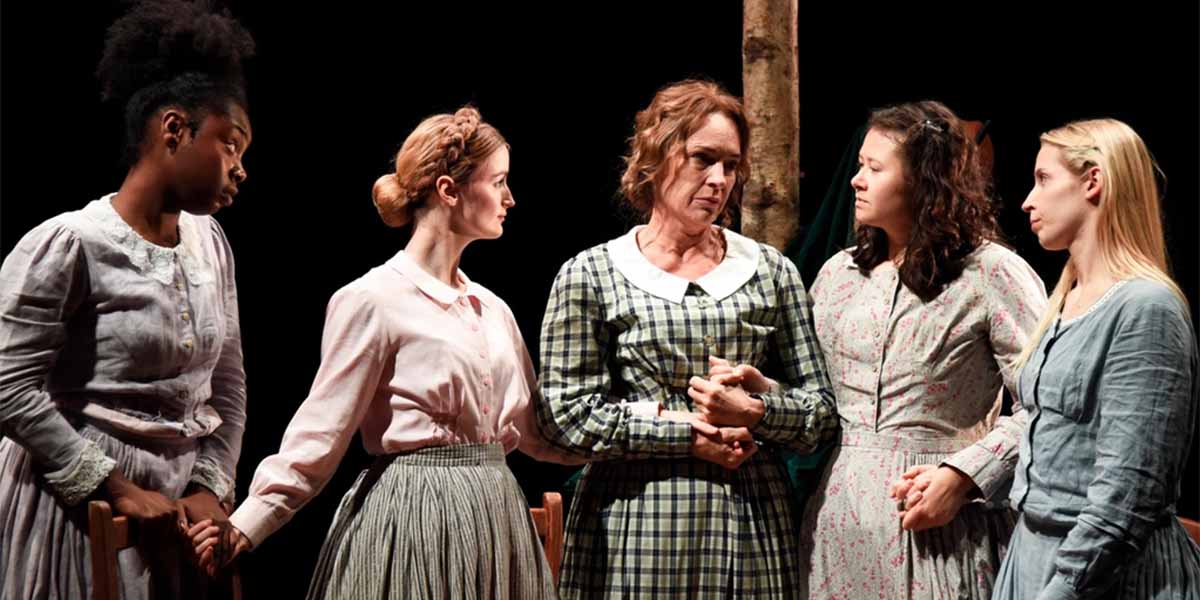 Five women in period costumes stand together, expressing emotion, with a tree in the background.